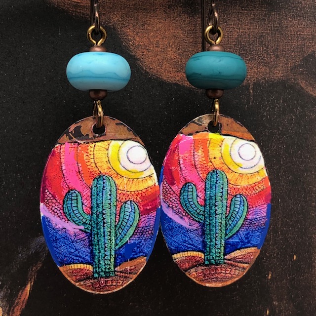 Boucles d'oreilles asymétriques composées de pendentifs en cuivre émaillé représentant des cactus verts sur un fond multicolore. Pièces uniques.