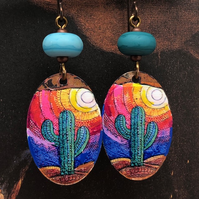 Boucles d'oreilles asymétriques composées de pendentifs en cuivre émaillé représentant des cactus verts sur un fond multicolore. Pièces uniques.