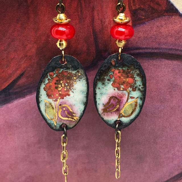 Boucles d'oreilles bohème composées de pendentifs en cuivre émaillé représentant une fleur rouge et un oiseau mauve, sur un décor parsemé de touches d'or.