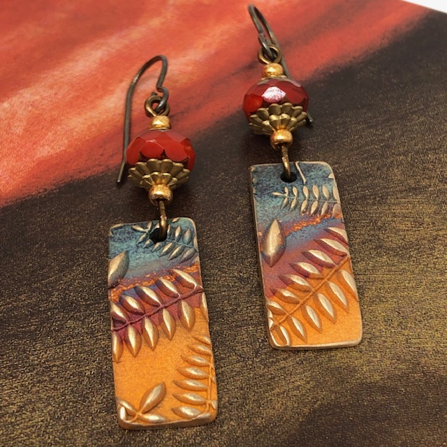 Boucles d'oreilles bohème composées de pendentifs artisanaux en bronze représentant des fougères dorées sur un fond multicolore. Pièces uniques.