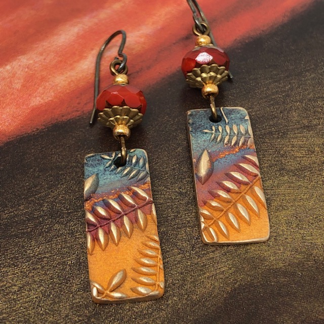 Boucles d'oreilles bohème composées de pendentifs artisanaux en bronze représentant des fougères dorées sur un fond multicolore. Pièces uniques.