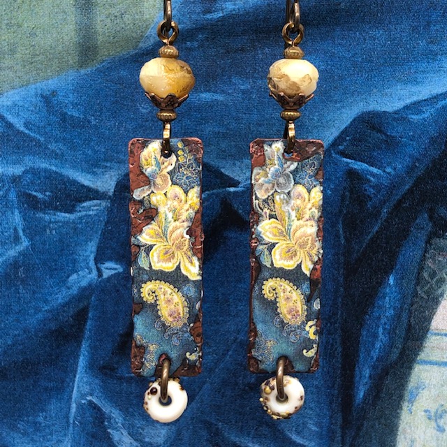 Boucles d'oreilles bohèmes et asymétriques composées de pendentifs en cuivre émaillé qui représentent des iris jaunes sur un fond bleu foncé.