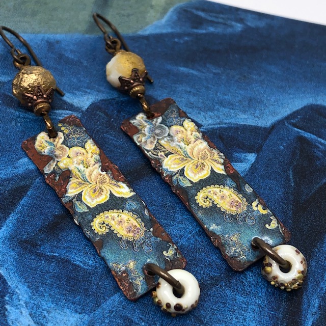 Boucles d'oreilles bohèmes et asymétriques composées de pendentifs en cuivre émaillé qui représentent des iris jaunes sur un fond bleu foncé.
