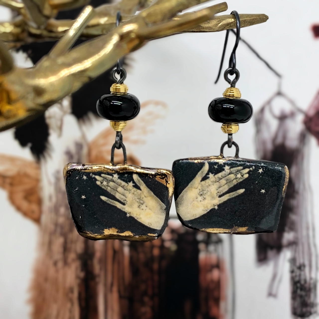 Boucles d'oreilles noir et or composées de pendentifs artisanaux en céramique qui représentent deux mains ouvertes sous une pluie d'or. Modèle unique.