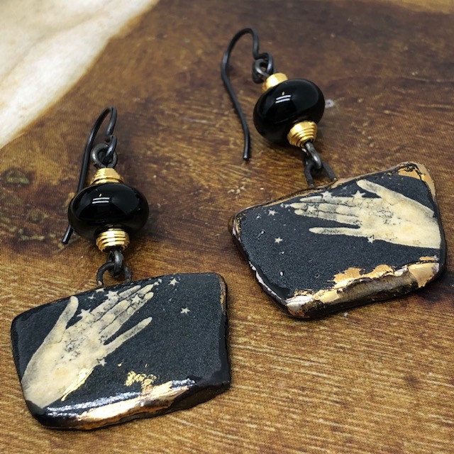 Boucles d'oreilles noir et or composées de pendentifs artisanaux en céramique qui représentent deux mains ouvertes sous une pluie d'or. Modèle unique.