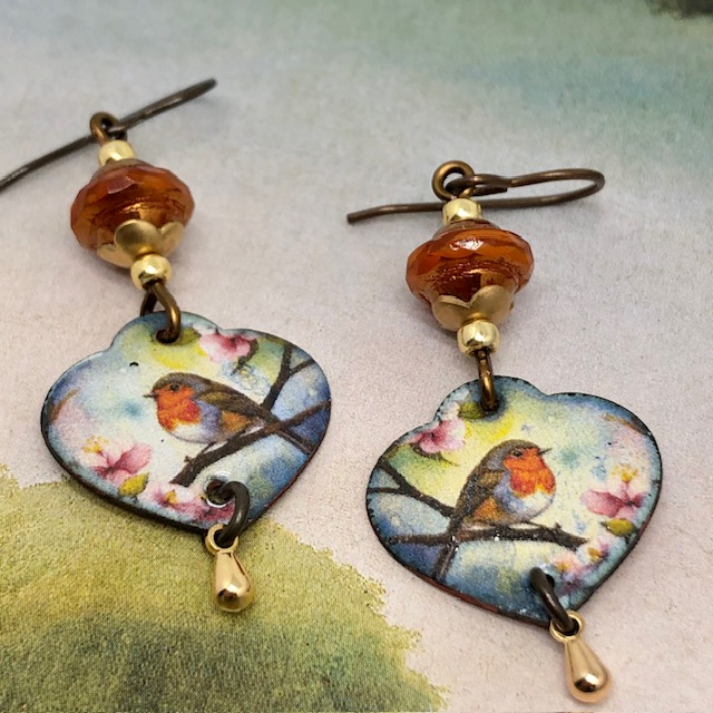 Boucles d’oreilles poétiques composées de pendentifs en cuivre émaillé en forme de coeur avec un rouge-gorge sur un fond multicolore. Bijoux uniques.