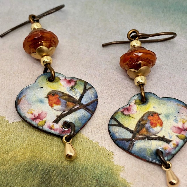 Boucles d’oreilles poétiques composées de pendentifs en cuivre émaillé en forme de coeur avec un rouge-gorge sur un fond multicolore. Bijoux uniques.