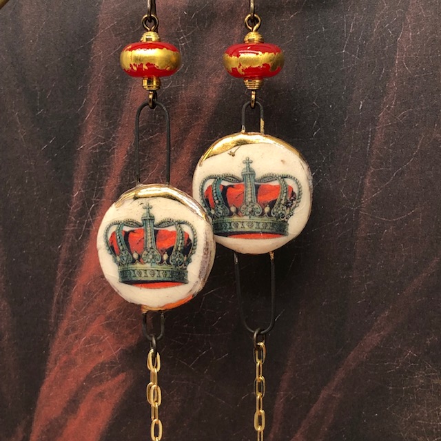 Longues boucles d'oreilles asymétriques composées de pendentifs artisanaux en céramique représentant une couronne posé sur un coussin rouge. Pièces uniques.