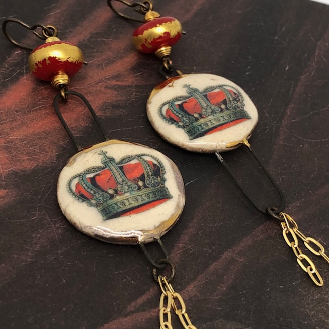 Longues boucles d'oreilles asymétriques composées de pendentifs artisanaux en céramique représentant une couronne posé sur un coussin rouge. Pièces uniques.