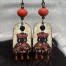 Boucles d’oreilles poétiques et composées de pendentifs en cuivre émaillé avec le portrait d'une femme vêtue d'une robe noire ornée de feuilles d'automne.