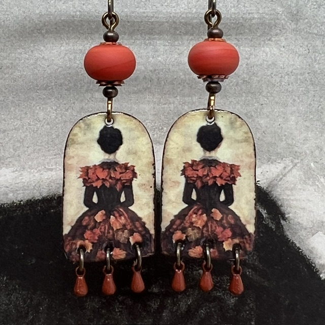 Boucles d’oreilles poétiques et composées de pendentifs en cuivre émaillé avec le portrait d'une femme vêtue d'une robe noire ornée de feuilles d'automne.