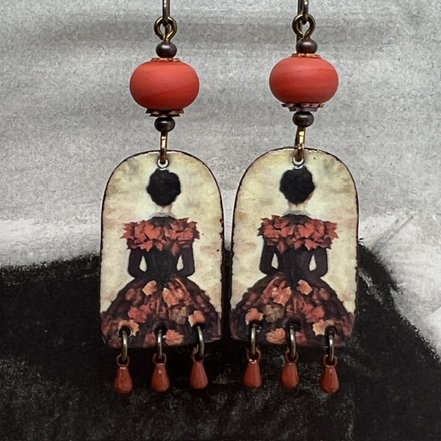 Boucles d’oreilles poétiques et composées de pendentifs en cuivre émaillé avec le portrait d'une femme vêtue d'une robe noire ornée de feuilles d'automne.