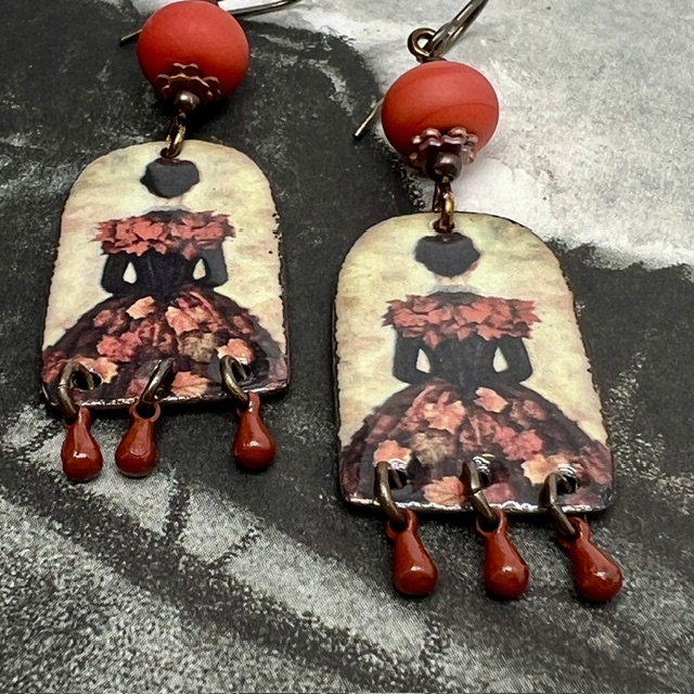 Boucles d’oreilles poétiques et composées de pendentifs en cuivre émaillé avec le portrait d'une femme vêtue d'une robe noire ornée de feuilles d'automne.