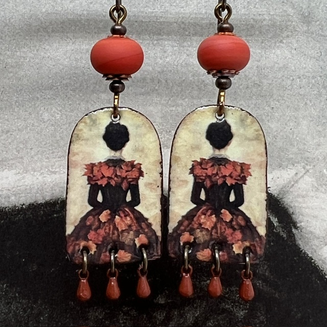 Boucles d’oreilles poétiques et composées de pendentifs en cuivre émaillé avec le portrait d'une femme vêtue d'une robe noire ornée de feuilles d'automne.