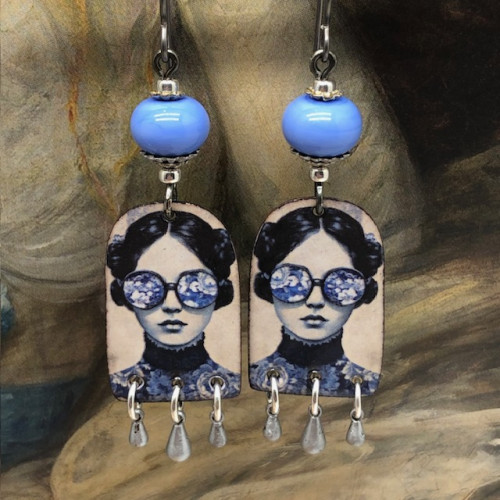 Boucles d'oreilles poétiques composées de pendentifs en cuivre émaillé avec le portrait d'une femme portant des lunettes décorées de fleurs bleu ciel.