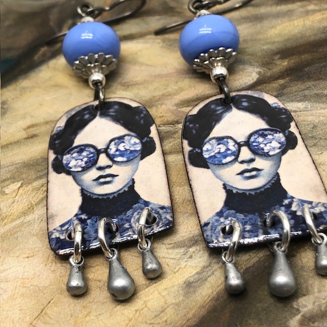 Boucles d'oreilles poétiques composées de pendentifs en cuivre émaillé avec le portrait d'une femme portant des lunettes décorées de fleurs bleu ciel.