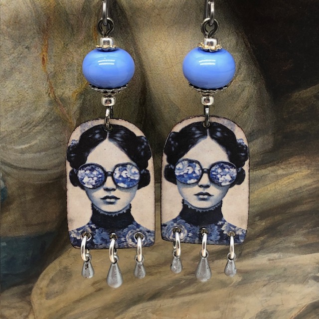 Boucles d'oreilles poétiques composées de pendentifs en cuivre émaillé avec le portrait d'une femme portant des lunettes décorées de fleurs bleu ciel.