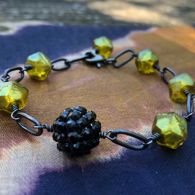 Bracelet composé de perles en verre tchèque et d'une perle noire en plasticine ornée de strass. Chaîne et fermoir en laiton. Pièce unique.